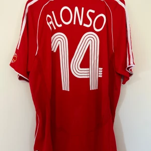 Liverpool fotbollströja med Alonso 14 - Säljer en röd Liverpool fotbollströja från Adidas med Alonso och nummer 14 på ryggen. Tröjan har korta ärmar och det klassiska Carlsberg-trycket på framsidan. Den är i bra skick och perfekt för alla Liverpool-fans!