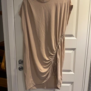 Beige tunika från Gina Tricot - Säljer en stilren beige tunika från Gina Tricot. Tunika har en snygg drapering med snörning på sidan och är ärmlös. Perfekt för en avslappnad look.