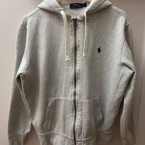 Grå hoodie från Ralph Lauren - Säljer en stilren grå hoodie från Ralph Lauren med dragkedja och justerbar huva. Den har en broderad logotyp på bröstet och praktiska fickor framtill. Perfekt för en avslappnad look.