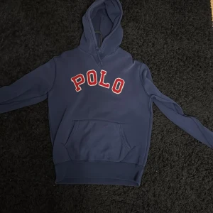 Blå hoodie från Ralph Lauren - Säljer en snygg blå hoodie från Ralph Lauren med texten 'POLO' i rött och vitt på framsidan. Tröjan har en klassisk design med huva och en stor ficka framtill. Perfekt för en avslappnad stil.