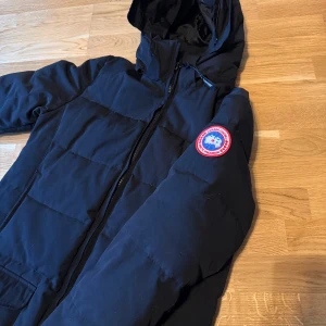 Svart dunjacka med pälsdetalj - Säljer en svart dunjacka från Canada Goose med pälsdetalj på huvan. Jackan har en dragkedja framtill och en ikonisk logga på ärmen. Perfekt för kalla vinterdagar med sin varma och skyddande design. passar även M men är storlek S. har inga tydliga defekter jag märkt av eller som syns