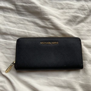 Svart plånbok från Michael Kors - Snygg svart plånbok från Michael Kors med guldfärgad dragkedja och logotyp. Perfekt för att hålla ordning på kort och kontanter med flera fack inuti. Stilren design som passar till det mesta. Defekter finns se bild 3,4