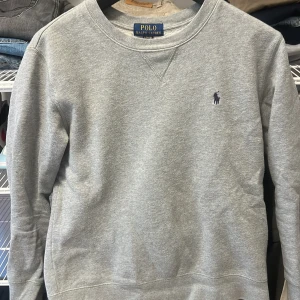 Grå sweatshirt från Ralph Lauren - Säljer en klassisk grå sweatshirt från Ralph Lauren med det ikoniska broderade logotypen på bröstet. Tröjan har rund hals och långa ärmar, perfekt för en avslappnad stil.