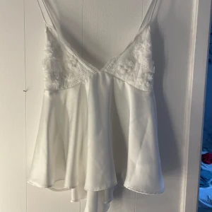 Vit satinlinne med spetsdetaljer - Elegant vit satinlinne med tunna axelband och v-ringning. Toppen har vackra spetsdetaljer på bysten och en knytning i ryggen för en fin touch. Perfekt för en stilren look. Använd 1 gång 