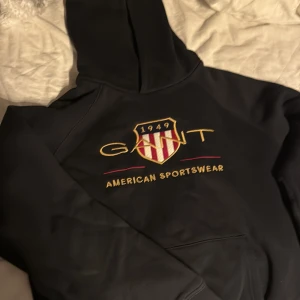 Svart hoodie från GANT - Säljer en svart hoodie från GANT med broderad logga och texten 'American Sportswear' på framsidan. Hoodien har en klassisk design med känguruficka och justerbar huva. Perfekt för en avslappnad stil.
