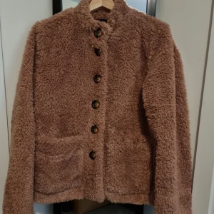 Brun teddyjacka från Object - Mysig brun teddyjacka från Object med knappar framtill och två fickor. Perfekt för kyliga dagar när du vill hålla dig varm och stilren. Jackan har en klassisk passform. Den har aldrig blivit använd. 