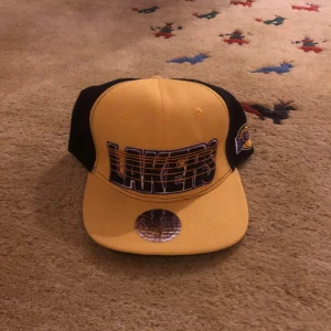 Gul och svart keps med Lakers-logga - Snygg gul och svart keps med Lakers-logga framtill. Kepsen har en platt skärm och justerbar passform. Perfekt för sportiga tillfällen eller som en stilren accessoar.