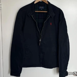 Svart jacka från Polo Ralph Lauren - Snygg svart jacka från Polo Ralph Lauren med dragkedja och röd broderad logga på bröstet. Jackan har en klassisk krage och är perfekt för en stilren look. Den har ett rutigt foder som ger en extra touch av stil.