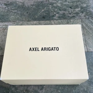 Beige sneakers från Axel Arigato - Snygga beige sneakers från Axel Arigato med hög design och vita snören. Skorna har en stilren look med perforerade detaljer och en robust sula. Perfekta för en trendig och avslappnad stil. Nypris 3500kr