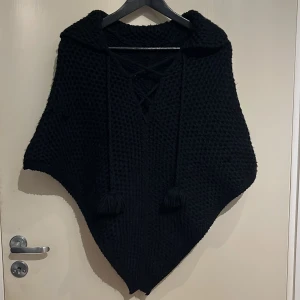 Svart stickad poncho med snörning - Säljer en svart stickad poncho med snygg snörning framtill. Den har en mysig och lös passform, perfekt för kyliga dagar. Ponchon är utan ärmar och har dekorativa tofsar som ger en extra touch. Perfekt för att hålla sig varm och stilren.