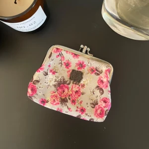 Blommig plånbok med metallspänne - Charmig liten plånbok med ett blommigt mönster i rosa och beige toner. Den har ett klassiskt metallspänne som ger en vintagekänsla. Perfekt för att hålla ordning på mynt och småsaker. 