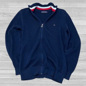 Tommy Hilfiger zip - Snygg mörkblå tröja från Tommy Hilfiger med dragkedja och ribbad krage. Kragen har ränder i rött, vitt och blått. Perfekt för en stilren look. Tröjan är välbehållen och har inga skador eller liknande.