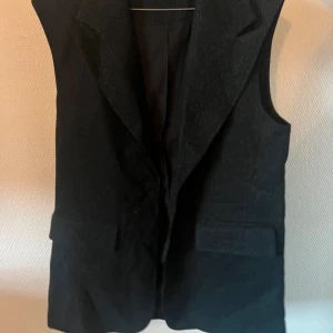 Svart väst med diskreta ränder - Snygg svart väst med diskreta ränder och klassisk krage. Perfekt för en stilren look. Västen har knappar framtill och två fickor. Ett mångsidigt plagg för olika tillfällen.