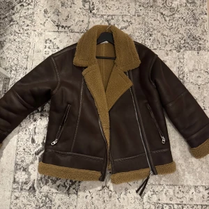 Brun skinnjacka med teddyfoder - Säljer en snygg brun skinnjacka med mysigt teddyfoder. Jackan har en klassisk design med dragkedja och två fickor framtill. Perfekt för kyliga dagar med sin varma och stiliga look. Jackan har en liten skada nertill vid dragkedjan, inget som märks av. Köpt för ca 2 år sedan, däremot använd ett fåtal gånger. Jackan är från Zara