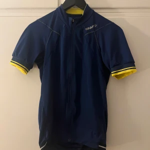 Blå cykeltröja från Craft - Säljer en blå cykeltröja från Craft med korta ärmar och dragkedja framtill. Tröjan har gula detaljer vid ärmslut och krage samt praktiska fickor på baksidan. Perfekt för cykling och andra sportaktiviteter.