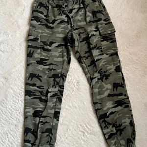 Camouflage cargobyxor - Snygga cargobyxor i camouflage från MISU. Byxorna har en elastisk midja och praktiska fickor på sidorna. Perfekta för en avslappnad stil med en cool edge.