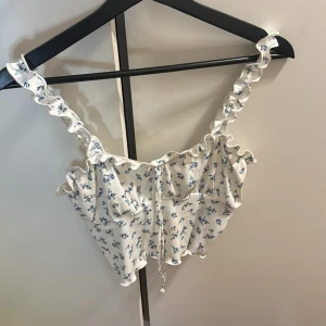 Vit blommig topp med volangdetaljer - Söt vit topp med blå blommönster och volangdetaljer. Toppen har smala axelband och en knytning framtill som ger en romantisk touch. Perfekt för varma sommardagar. Den är från SHEIN