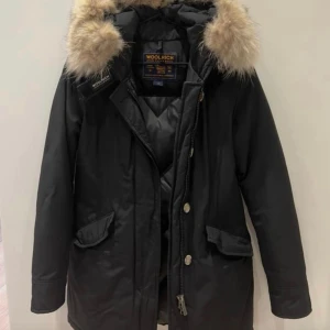 Svart parkajacka från Woolrich - Säljer en svart parkajacka från Woolrich med en mysig pälsfodrad huva. Jackan har dragkedja och knappar framtill samt praktiska fickor. Perfekt för kyliga dagar.