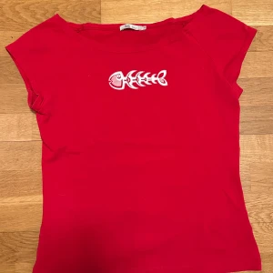 Röd kortärmad topp med fiskbenstryck - Säljer en röd kortärmad topp från Desigual med ett unikt fiskbenstryck i vitt på framsidan. Perfekt för en avslappnad stil. Toppen har en rund halsringning och är tillverkad i ett mjukt material.