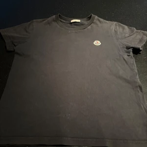 Svart t-shirt från Moncler - Säljer en klassisk svart t-shirt från Moncler med deras ikoniska logga broderad på bröstet. Perfekt för en stilren look. T-shirten har en rund halsringning och korta ärmar. Den har ett pytte litet hål men man ser ej. Den är för barn kanske 11-12 år gammalt.