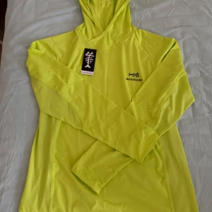 Neongul fishing hoodie från Bassdash - Säljer en neongul hoodie från Bassdash med UPF 50+ UV-skydd. Tillverkad i 84% polyester och 16% spandex, perfekt för utomhusaktiviteter. Tröjan har en enkel design med en liten logotyp på bröstet och ryggen.