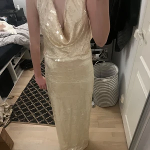 Champagnefärgad långklänning - Elegant champagnefärgad långklänning med djup v-ringning och öppen rygg. Klänningen har en glittrig yta som ger en lyxig känsla. Perfekt för speciella tillfällen.