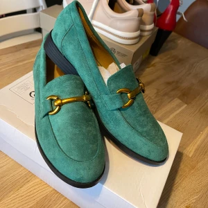 Gröna mockaloafers med gulddetaljer - Snygga gröna loafers  med guldfärgade detaljer på ovansidan. Skorna har en klassisk design med låg klack och rundad tå, perfekt för en stilren look.Helt nya