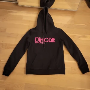 Svart hoodie från Dirt Cult - Säljer en svart hoodie från Dirt Cult med rosa tryck 'San Diego' på framsidan. Tröjan har en klassisk design med huva och långa ärmarHör av dig vid intresse!