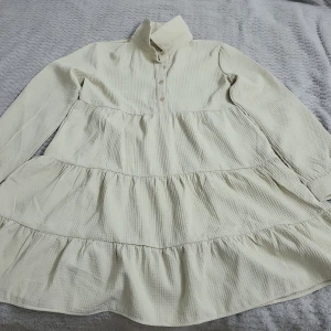 Beige klänning med volanger - Charmig beige klänning med volanger och långa ärmar. Den har en hög krage och knappar framtill. Perfekt för en stilren och bekväm look.