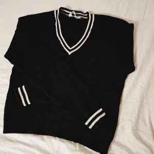 Svart stickad tröja från H&M - Säljer en svart stickad tröja från H&M med vita ränder på v-ringningen och ärmsluten. Tröjan har en klassisk design med ribbade detaljer och långa ärmar. Perfekt för en stilren look.