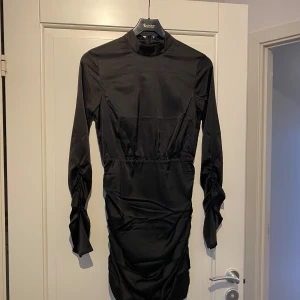Svart klänning med rynkdetaljer - Snygg svart klänning med långa ärmar och rynkdetaljer längs sidorna. Klänningen har en hög hals och är tillverkad i ett glansigt material som ger en elegant look. Perfekt för en kväll ute eller en speciell tillställning.