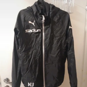 Svart vindjacka från Puma - Säljer en svart vindjacka från Puma med vit dragkedja och logga. Jackan har långa ärmar och en huva, perfekt för blåsiga dagar. Den är tillverkad i polyester och har en stilren design med Stadium-tryck på bröstet.