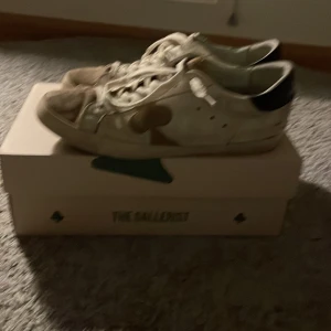 Vita sneakers från The Gallerist - Säljer ett par vita sneakers från The Gallerist med svarta detaljer på hälen. Skorna har snörning och en stilren design. Perfekta för en avslappnad stil.