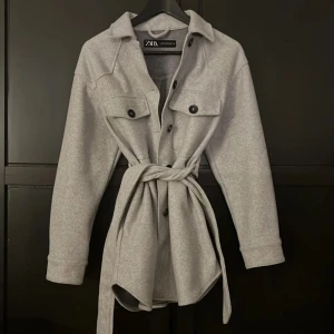 Kappa från Zara - Snygg grå overshirt från Zara med knappar och knytband i midjan. Den har långa ärmar och två bröstfickor med lock. Perfekt för lager-på-lager-stil. Har inte använt så mycket 