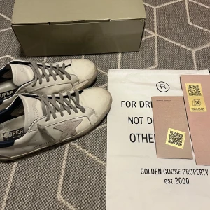Golden Goose sneakers - Säljer ett par Golden Goose sneakers i vitt med grå stjärndetalj och mörkblå häl. Skorna har en cool, sliten look och grå snörning. Perfekta för en avslappnad stil med en touch av lyx. Kommer med originalförpackning och påse. Priset kan diskuteras!