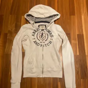 Säljer en vit zip up hoodie från Abercrombie & Fitch i bra skick. Den har en dragkedja framtill och ett snyggt tryck på bröstet. Insidan av huvan är randig i grått och vit. Tar emot trades annars är de fast pris🥰