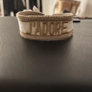 Beige armband med text - Säljer ett snyggt beige armband med texten 'J'ADORE' broderad i guld. Perfekt accessoar för att ge din outfit en personlig touch. Armbandet är justerbart och passar de flesta handleder. Perfekt för både vardag och fest!