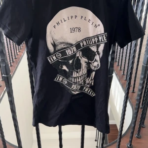 Svart t-shirt med dödskalle från Philipp Plein - Cool svart t-shirt från Philipp Plein Junior med ett stort dödskalletryck och texten '1978'. Perfekt för en edgy look! Passar tonåringar i storlek 14-15. T-shirten är i bra skick och redo för nya äventyr. 🖤