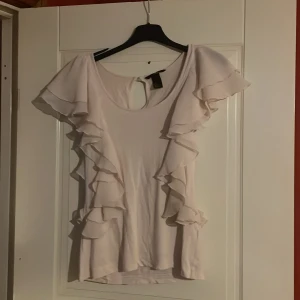 Beige topp med volanger från H&M - Säljer en superfin vit topp från H&M i storlek S. Den har volanger längs ärmarna och en rund halsringning med en liten öppning bak. Perfekt för sommarens fester eller en dag på stan. Toppen är i mycket bra skick och gjord av ett mjukt material.