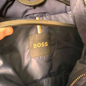 Mörkblå pufferjacka från BOSS (S M) - Säljer en snygg mörkblå pufferjacka från BOSS. Jackan har en dragkedja framtill och två praktiska fickor med dragkedjor. Den är perfekt för kalla dagar med sin vadderade design och huva. Passar både till vardags och lite finare tillfällen. 🧥