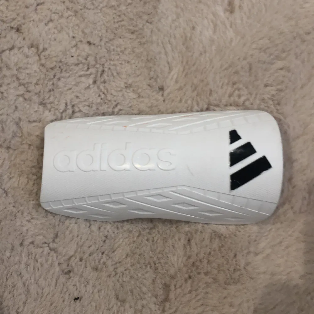 Säljer ett par vita benskydd från Adidas i mycket bra skick. De har en stilren design med svarta detaljer och Adidas-loggan. Perfekta för fotbollsträningen eller matchen. Passar bra för både kvinnor och män. Storleken är för alla. Sportswear.