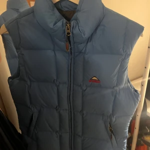 Blå dunväst från McKinley - Säljer en snygg blå dunväst från McKinley. Perfekt för kyliga dagar med sin quiltade design och dragkedja framtill. Västen har en hög krage för extra värme och stil. Passar bra för både vardag och utomhusaktiviteter. PRIS GÅR ATT DISKUTERA 