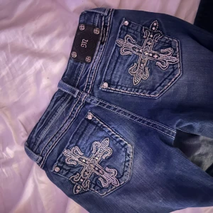 Blå jeans med broderade detaljer - Säljer ett par snygga blå jeans från Miss Me i storlek w28 L30. De har coola broderade korsdetaljer på bakfickorna. Jeansen är i bra skick och har en bootcut-stil och är låga i midjan. Helt nya som aldrig använts 
