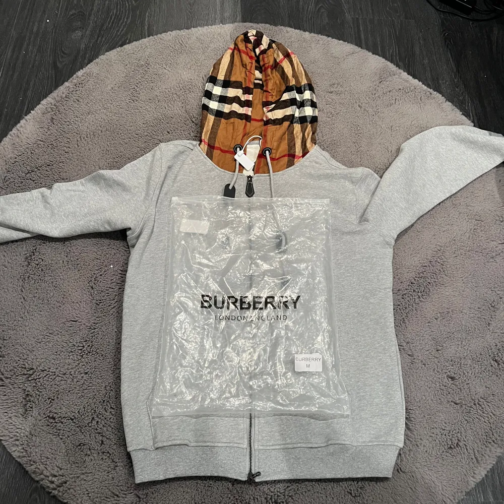 Säljer en snygg grå hoodie från Burberry med den ikoniska rutiga huvan i beige, svart och röd. Helt oanvänd perfekt för höst och vår! Den har dragkedja framtill och är i mycket bra skick. Passar både till vardags och lite finare tillfällen. 🧥✨. Tröjor & Koftor.