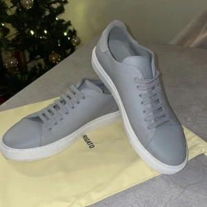 Grå sneakers från Axel Arigato - Säljer ett par stilrena grå sneakers från Axel Arigato i mycket bra skick. Skorna har en minimalistisk design med snörning och en vit sula. Nypris: 2450kr, i storlek 44!