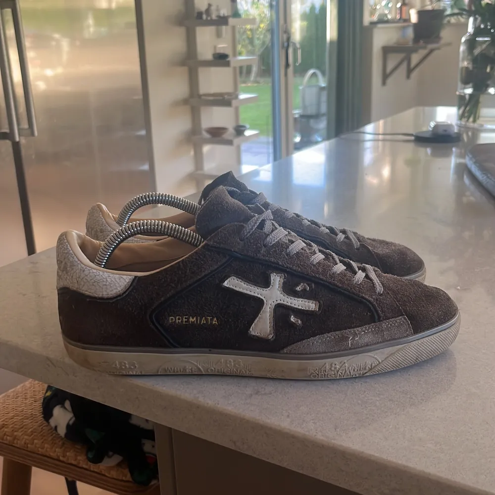 Säljer ett par snygga bruna sneakers från Premiata i mocka. De har en cool vit logga på sidan och grå snörning. Skorna är i bra skick med en stilren design som passar perfekt till vardags. Sulan har ett unikt mönster och är i gott skick. Perfekta för höst och vår! Tar gärna byten! . Kengät.