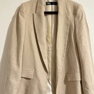 Beige kavaj från Zara - Säljer en stilren beige kavaj från Zara i mycket bra skick. Kavajen har en klassisk design med normal passform och långa ärmar. Perfekt för både vår och höst, och funkar lika bra till vardags som till fest. Materialet känns som en blandning av linne och bomull, vilket ger en skön och luftig känsla.