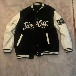 Svart och vit bomberjacka från H&M - Säljer en snygg svart och vit bomberjacka från H&M i storlek XS. Jackan har texten 'Playoffs' på framsidan och siffran '92' på ärmen. Den har långa ärmar och stängs med knappar.  Knappt använd och väldigt fin 