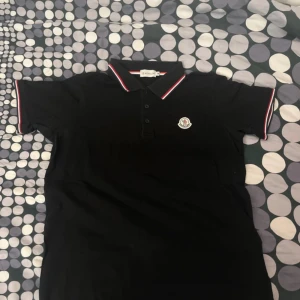 Svart pikétröja från Moncler - Säljer en snygg svart pikétröja från Moncler i mycket bra skick. Den har korta ärmar och en klassisk krage med röd och vit rand. Moncler-loggan är broderad på bröstet. Perfekt för både vardag och fest!