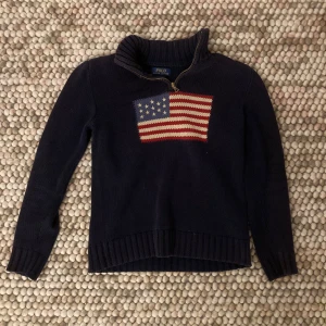 Mörkblå stickad tröja från Polo Ralph Lauren - Säljer en snygg mörkblå stickad tröja från Polo Ralph Lauren med en amerikansk flagga på bröstet. Tröjan har en dragkedja vid halsen och långa ärmar. Perfekt för kyliga dagar och ger en klassisk look. Passar både till vardags och lite finare tillfällen.